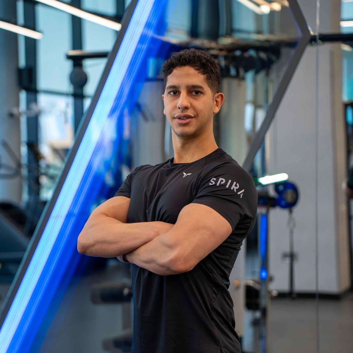 Abdulrahman Mekky with his arms crossed in a black Spira Club shirt, standing by a blue light strip on the gym floor